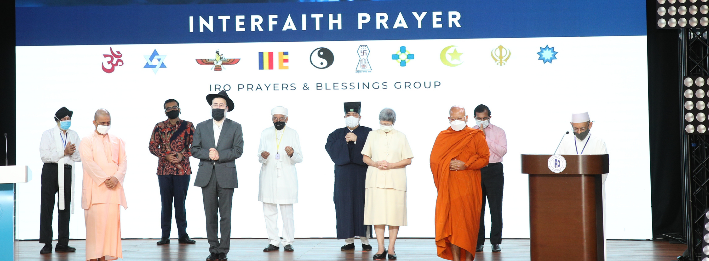 Diverse religious leaders stand on stage under "Interfaith Prayer" banner with world religion symbols.